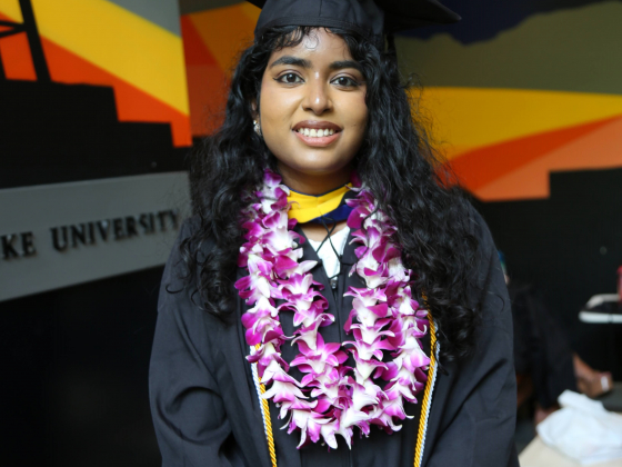 Portrait photo of sociology student Haripriya Dukkipati wearing graduation cap and gown and purple and white flower lei.