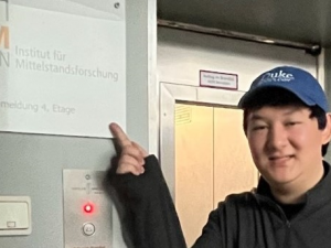 Photo of male student with Duke blue baseball cap on smiling and pointing at a sign that includes German language text