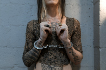 Photo of woman from neck to midriff with cuffs on wrist standing in front of white cinderblock wall with closed hands lifted in front of chest. 