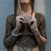 Photo of woman from neck to midriff with cuffs on wrist standing in front of white cinderblock wall with closed hands lifted in front of chest. 